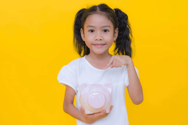 Asian girl holding piggy bank and pointing at it, promoting saving money and financial literacy for children against a vibrant yellow backdrop